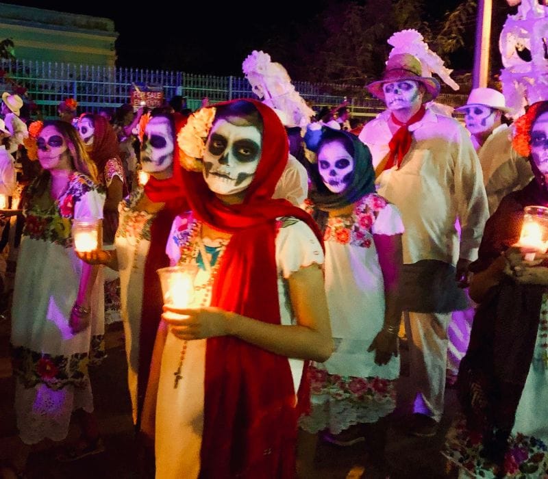 people with faces painted as skulls, holding a candle, walking in the paseo de animas parade in merida for hanal pixan